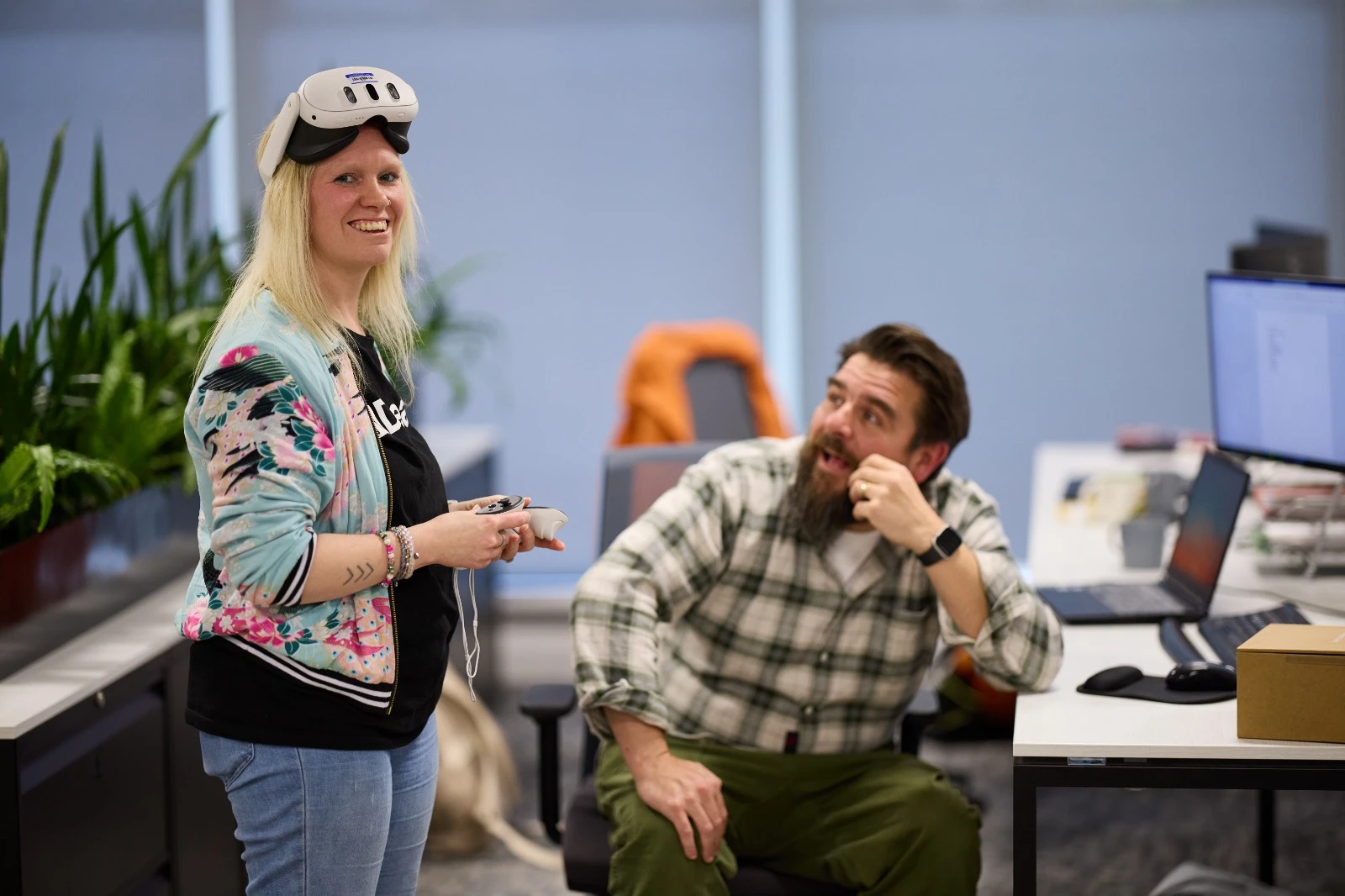 Ali Durrant wearing a raised VR headset, beside another person seated at a computer workstation.